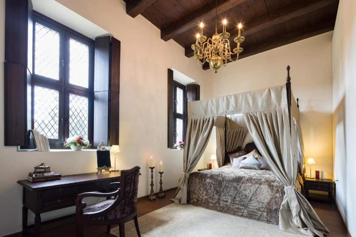 A bedroom with a four-poster bed, a desk with a chair, a chandelier, and a window with shutters.