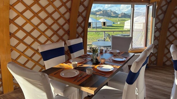 A dining area with a table set for four, a view of yurts outside, and a wooden lattice wall. - Khumug Tour Resort (Ulaanbaatar)