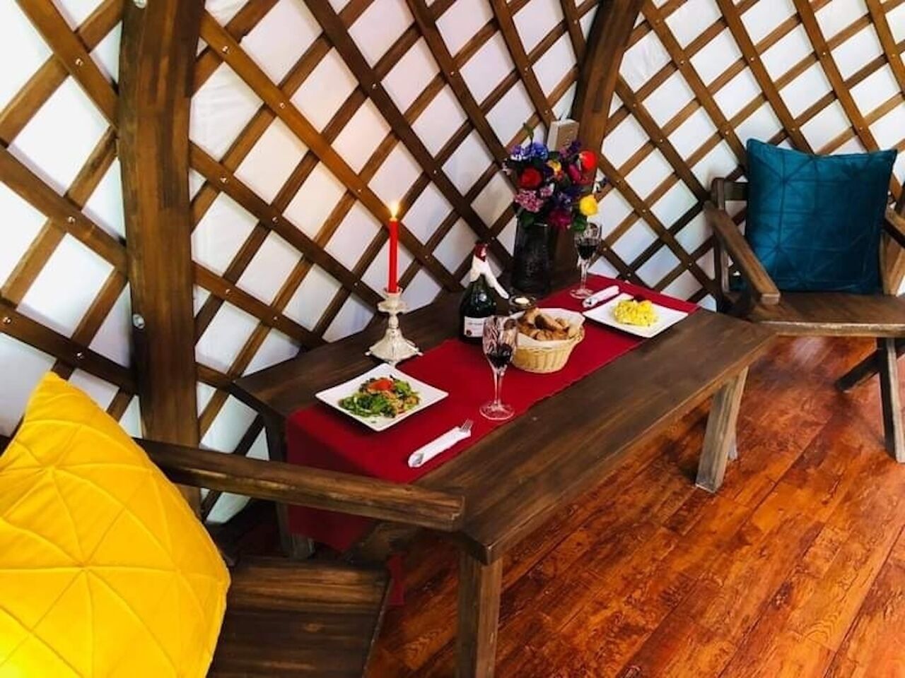 A dining area with a wooden table set for two, a red placemat, a candle, a bottle of wine, and a basket of bread.