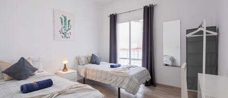 A bedroom with two beds, a bedside table with a lamp, a window with curtains, a mirror, and a wardrobe.