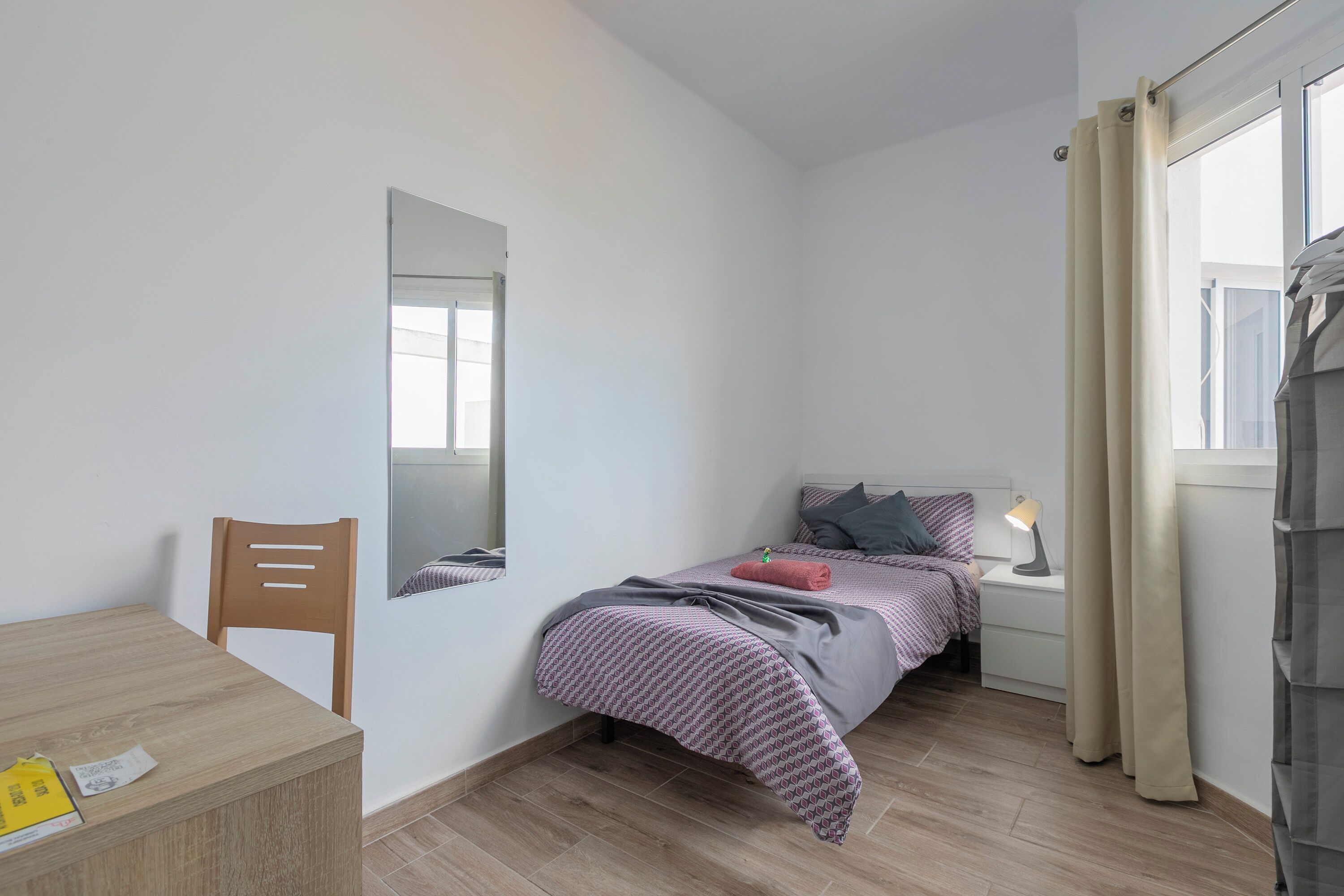 A bedroom with a bed, a bedside table with a lamp, a chair, a desk, and a window with curtains.