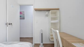 Une chambre à coucher avec un lit, un radiateur blanc, un banc en bois et une armoire blanche où sont suspendus des vêtements.
