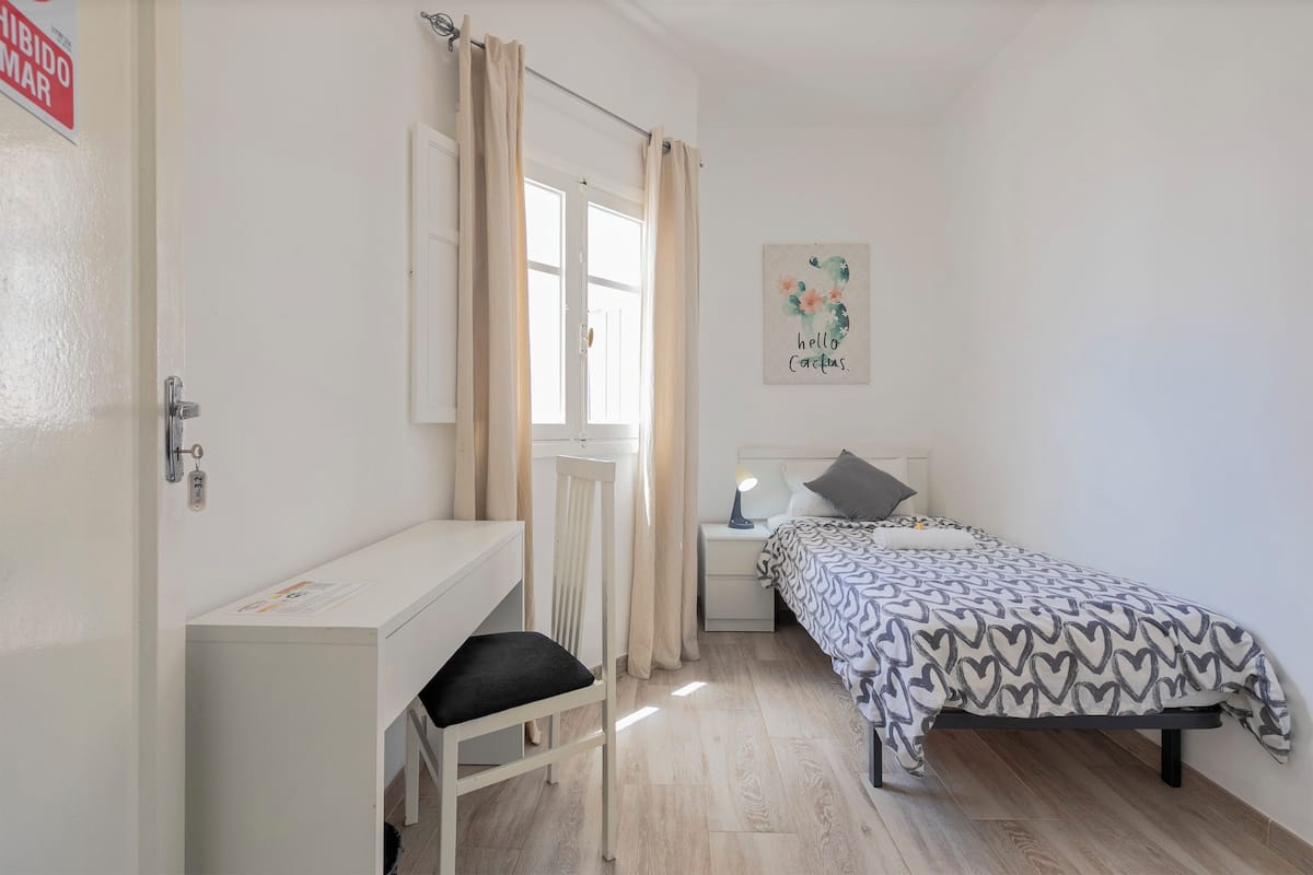 A bedroom with a bed, a desk, a chair, a nightstand, and a window with curtains.
