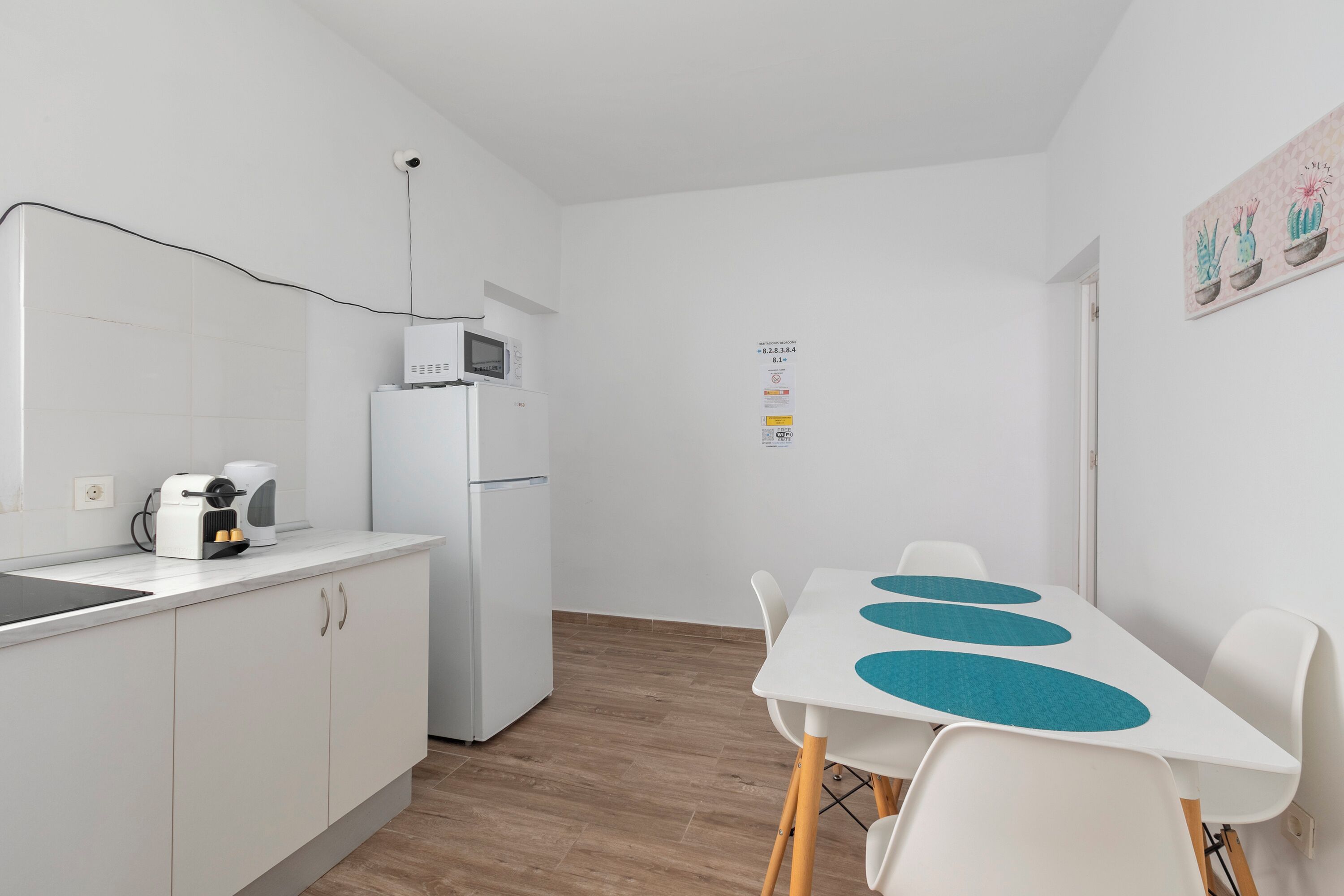 Une cuisine compacte équipée d’un four à micro-ondes, d’une machine à café et d’un réfrigérateur. On y trouve une table à manger avec des chaises, ainsi qu’une œuvre d’art représentant des cactus, accrochée au mur.