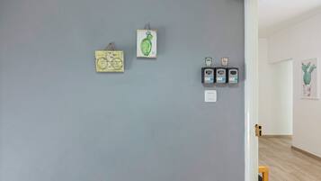 Quarto com parede cinzenta, porta de madeira e uma cadeira de madeira pequena. Há três quadros na parede: um de uma bicicleta, um de um cacto e três pequenos objetos pretos com números.