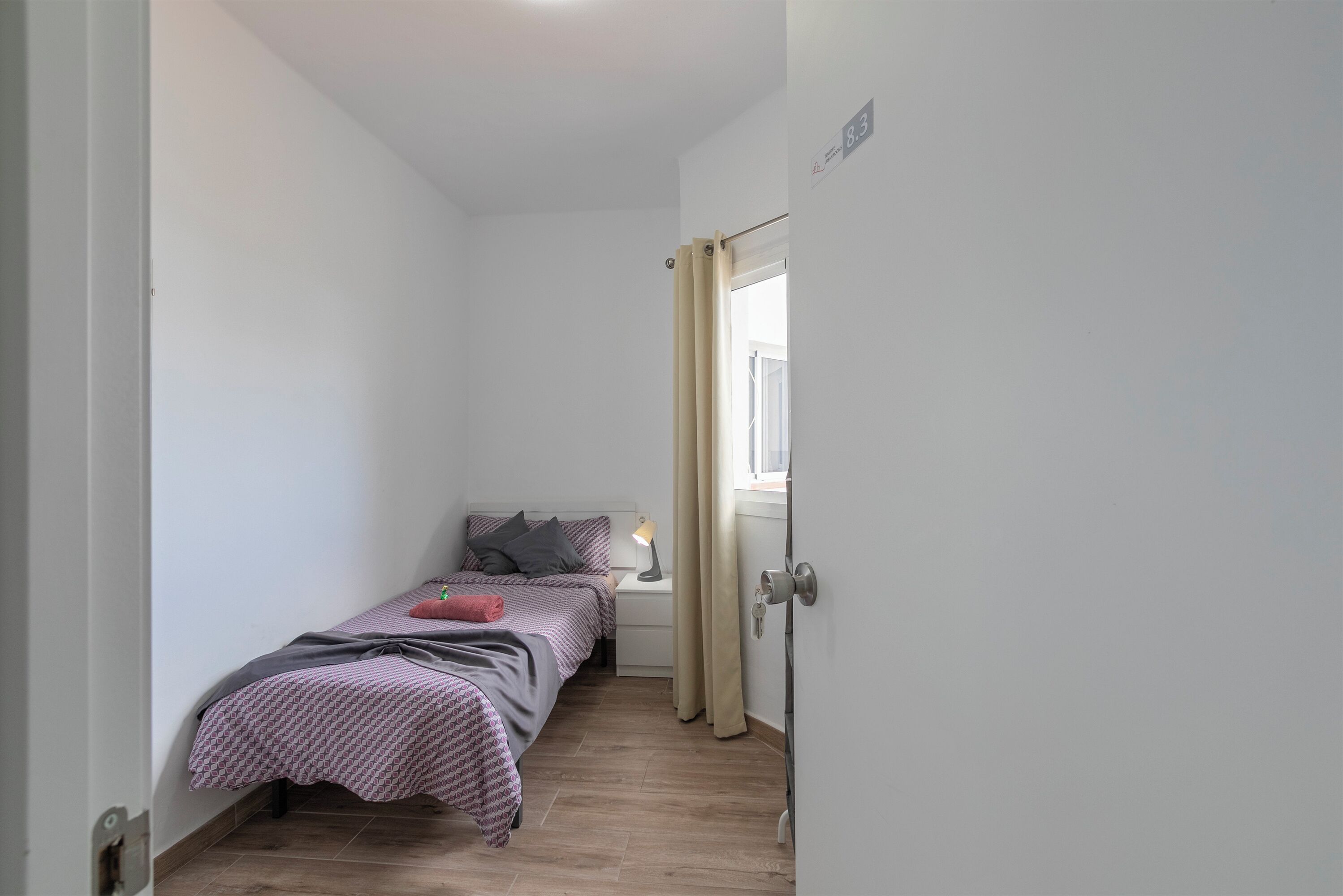 Een compacte, verzorgde slaapkamer met een enkel bed, nachtkastje, lamp en een raam met gordijnen.