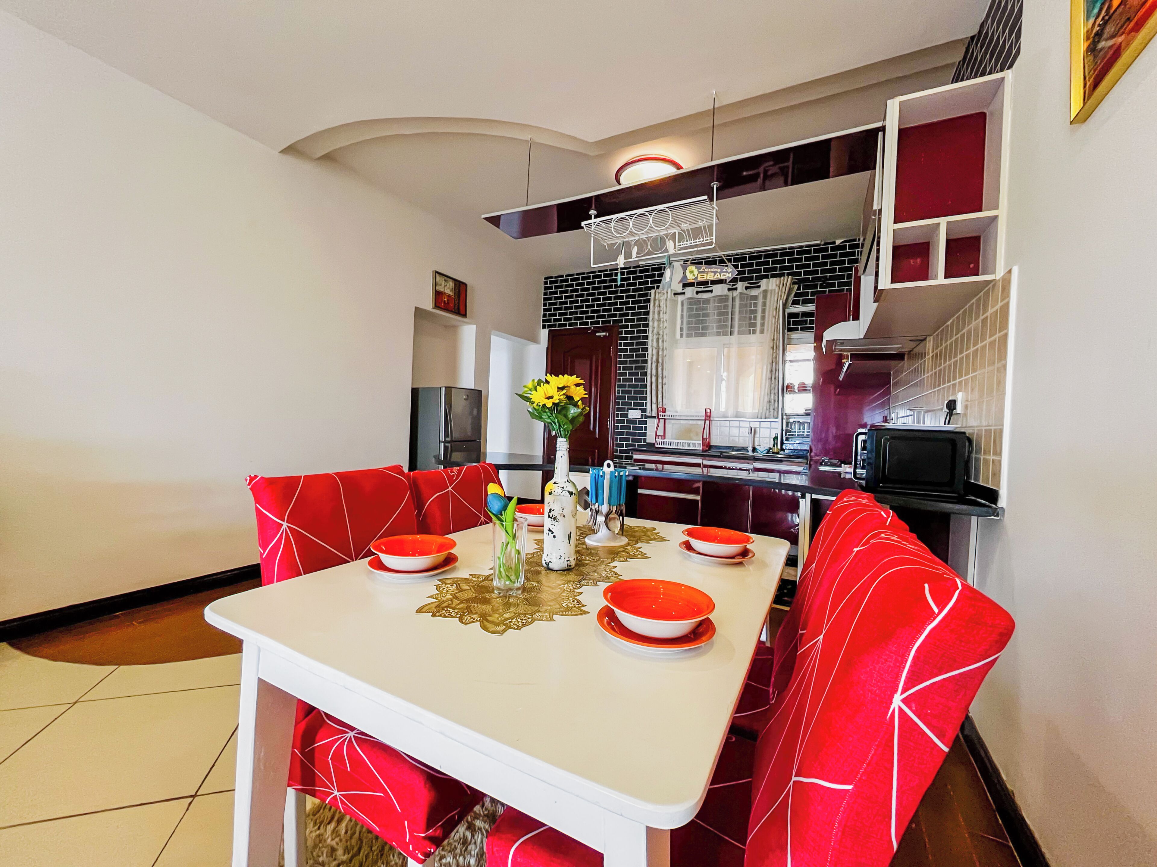 Un'area pranzo con un tavolo bianco e sedie rosse, un vaso con fiori e una cucina sullo sfondo.