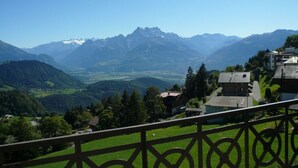 Un paisaje montañoso con una vista despejada de los Alpes al fondo. Se ve un edificio con un balcón en primer plano.