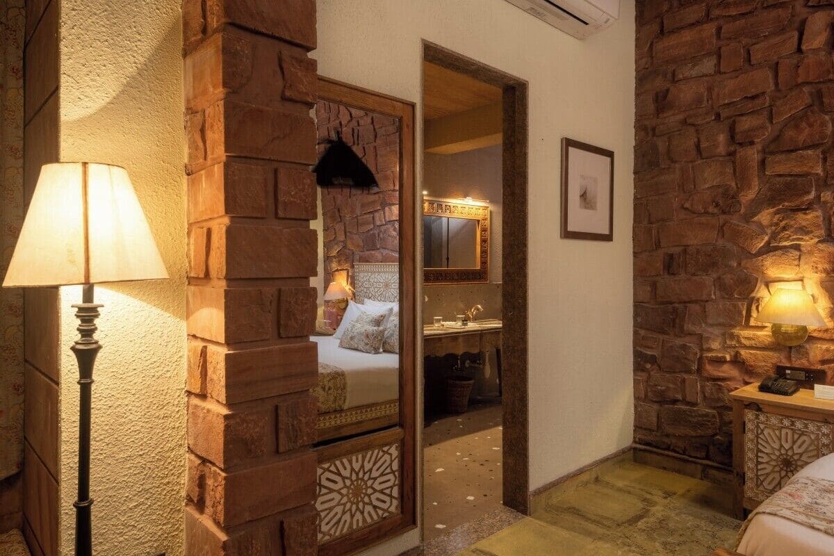 A cozy room with a stone wall, a bed with a patterned bedspread, a bedside table with a lamp, and a mirror on the wall.