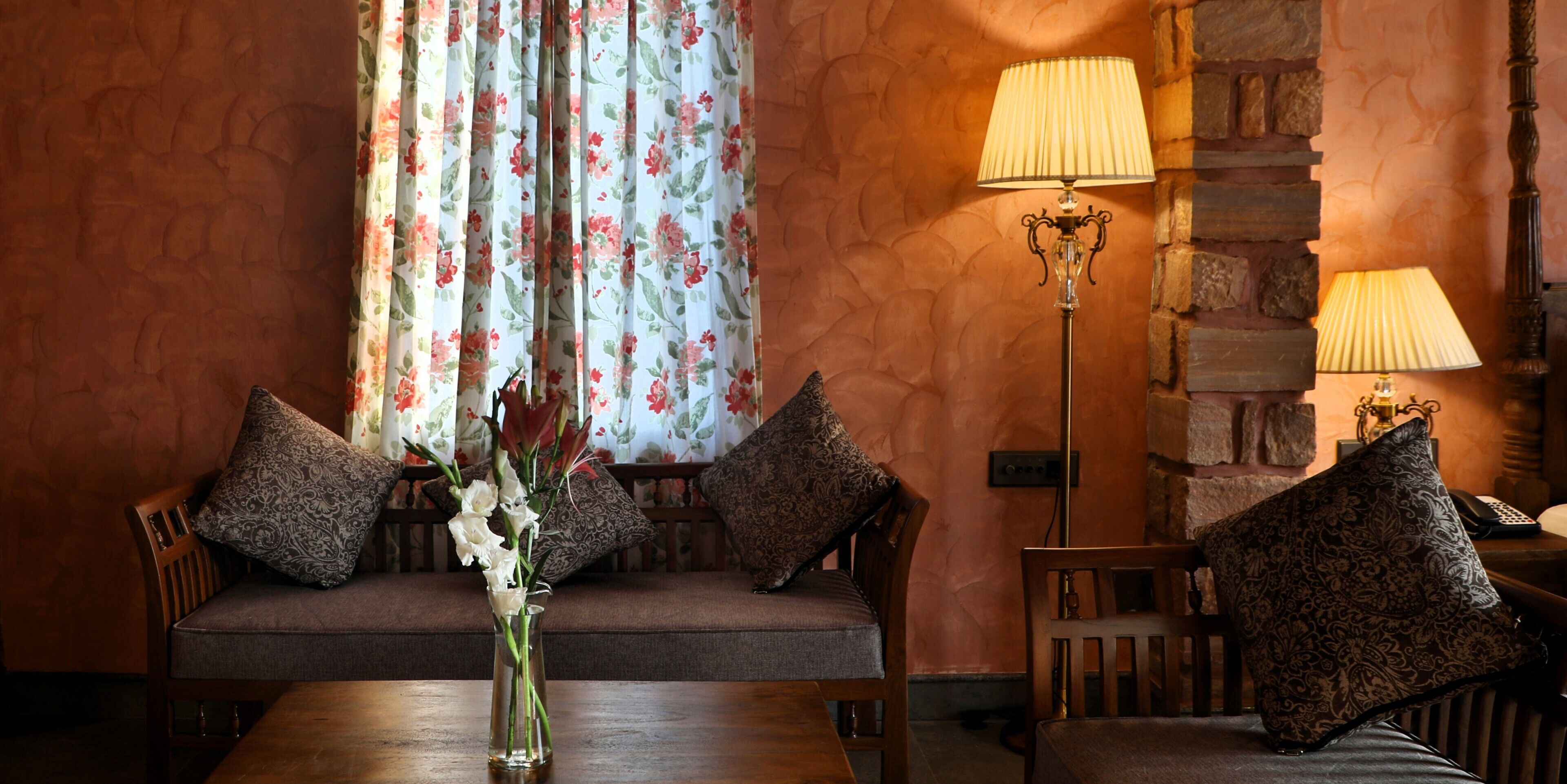 A room with a wooden table, two chairs, a vase with flowers, a sofa with patterned cushions, and two wall-mounted lamps.