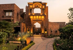 A stone building with an ornate archway, a pathway, and a garden with various plants. - Govind Bagh Resort (Jodhpur)