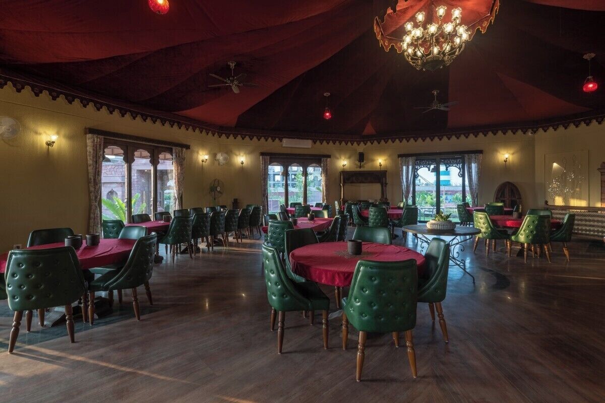 Un vaste espace dédié à la salle à manger, avec des tables rondes et des chaises recouvertes d’un tissu vert, de grandes fenêtres et un lustre.