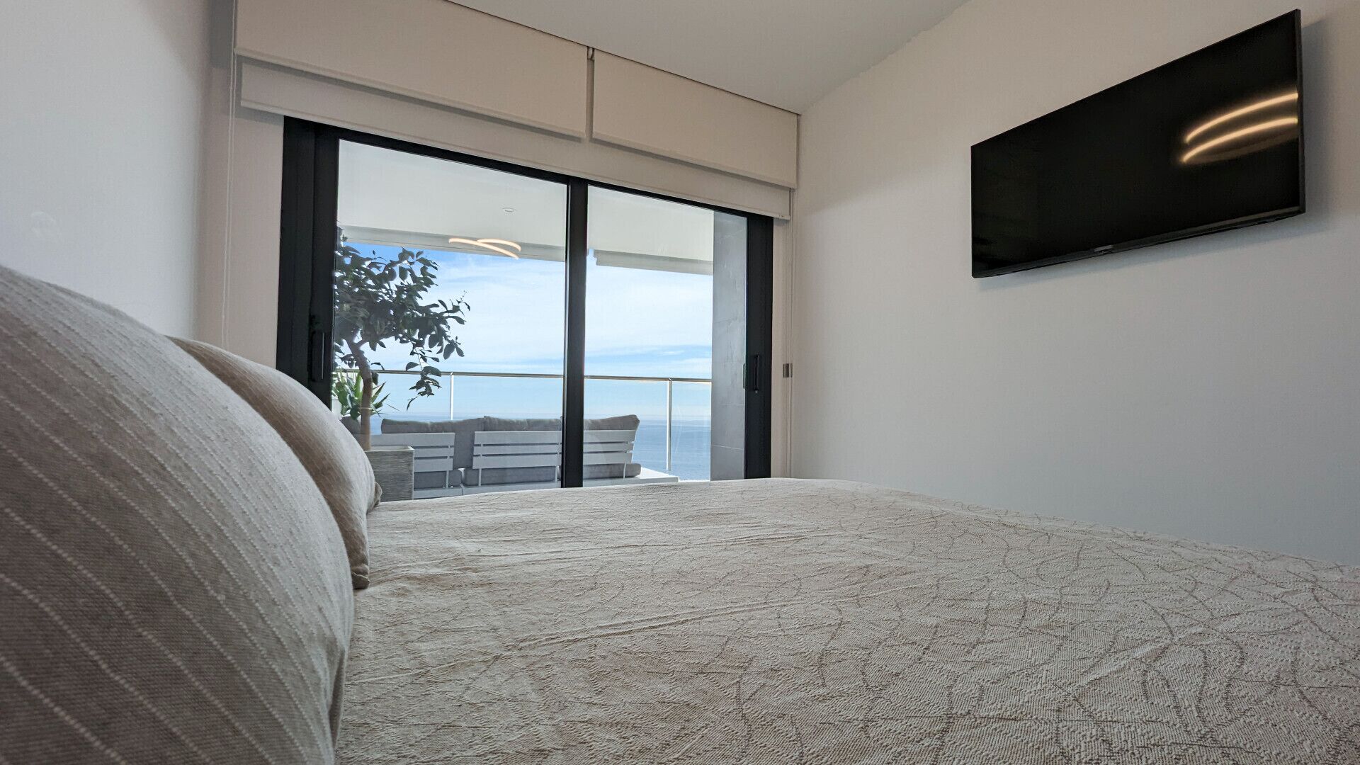 A bedroom with a large bed, a TV mounted on the wall, and a balcony with a view of the sea.