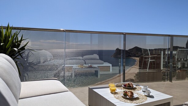 A modern balcony with a glass barrier, a white sofa, a small table with breakfast items, and a view of the sea and buildings. - SUNSET CLIFFS top apartment (Benidorm)
