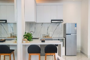 Une cuisine moderne avec des meubles blancs, un pare-feu en marbre, un réfrigérateur en acier inoxydable et deux chaises noires autour d’une table blanche.