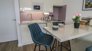 A modern kitchen with white cabinets, a built-in microwave, a sink, and a dining area with two chairs and a table. - Lan y Mor (Porthmadog)