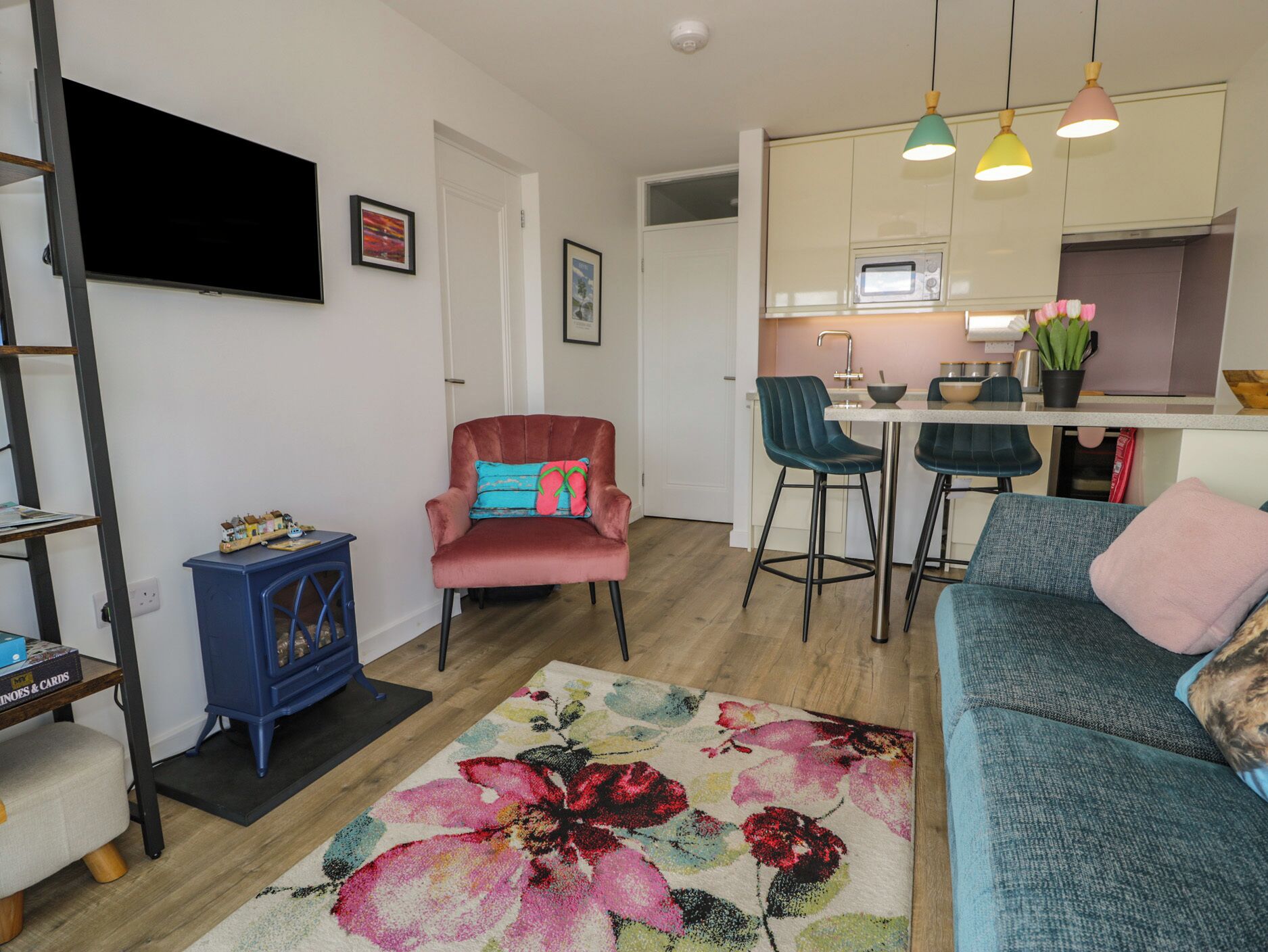 A modern living room with a sofa, a red armchair, a blue cabinet, a television, and a kitchen area with bar stools and pendant lights.