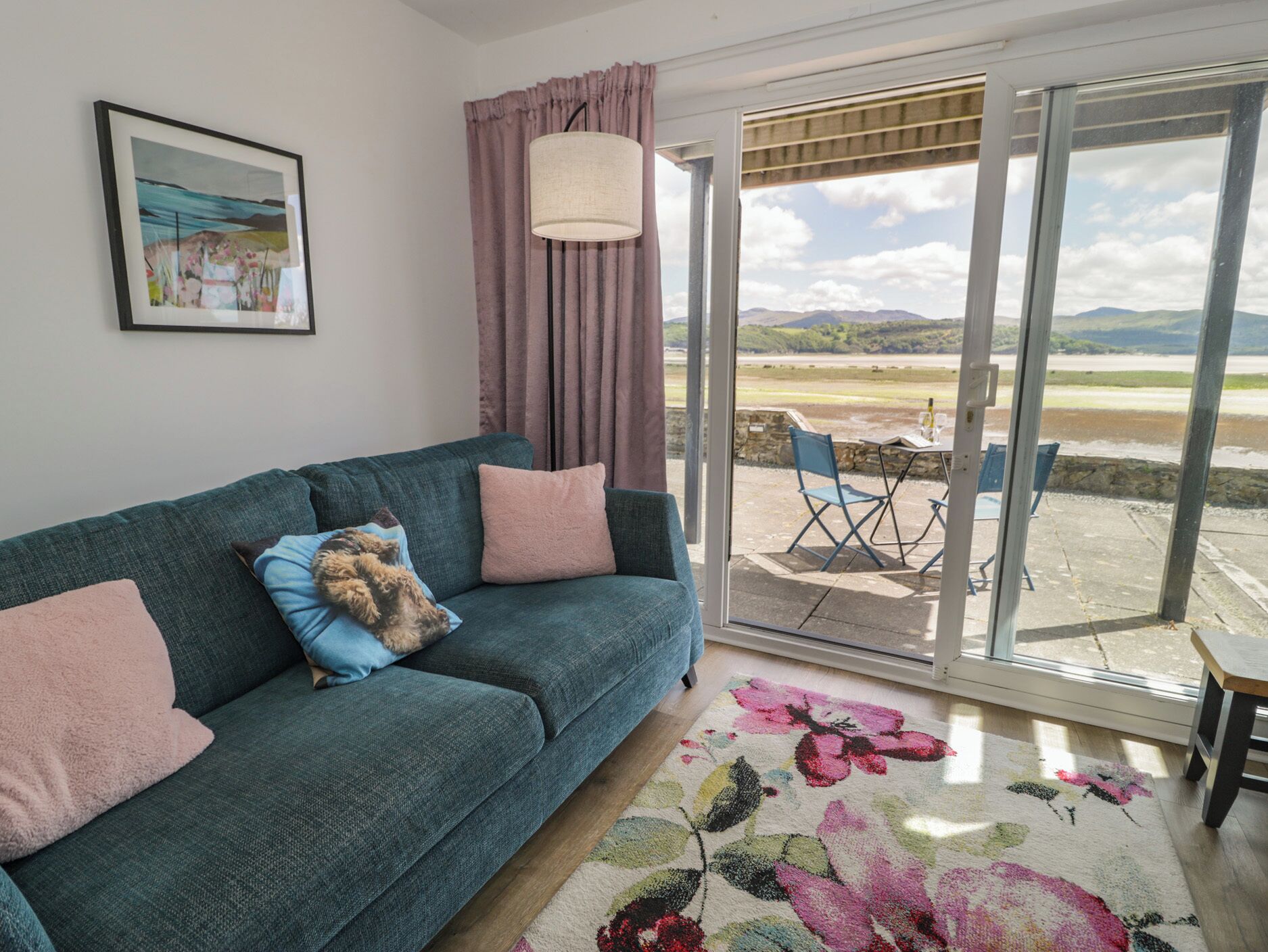 A living room with a sofa, a floral rug, a table with chairs, a large window with a view of the countryside, and a painting on the wall.