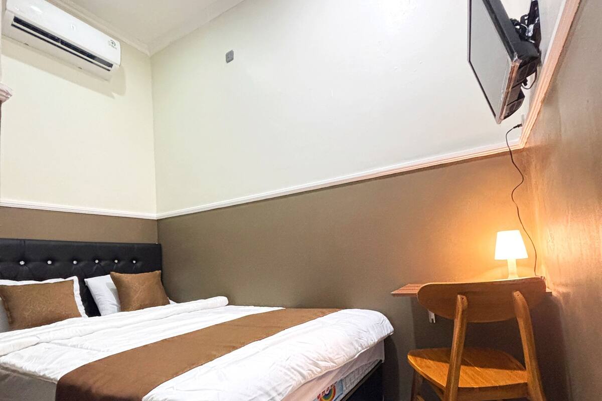 A bedroom with a bed, a chair, a desk, an air conditioner, and a wall-mounted TV.