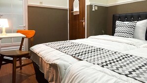 A hotel room with a bed, a desk with a chair, a window, and an air conditioning unit. - Griya Inn (Semarang)