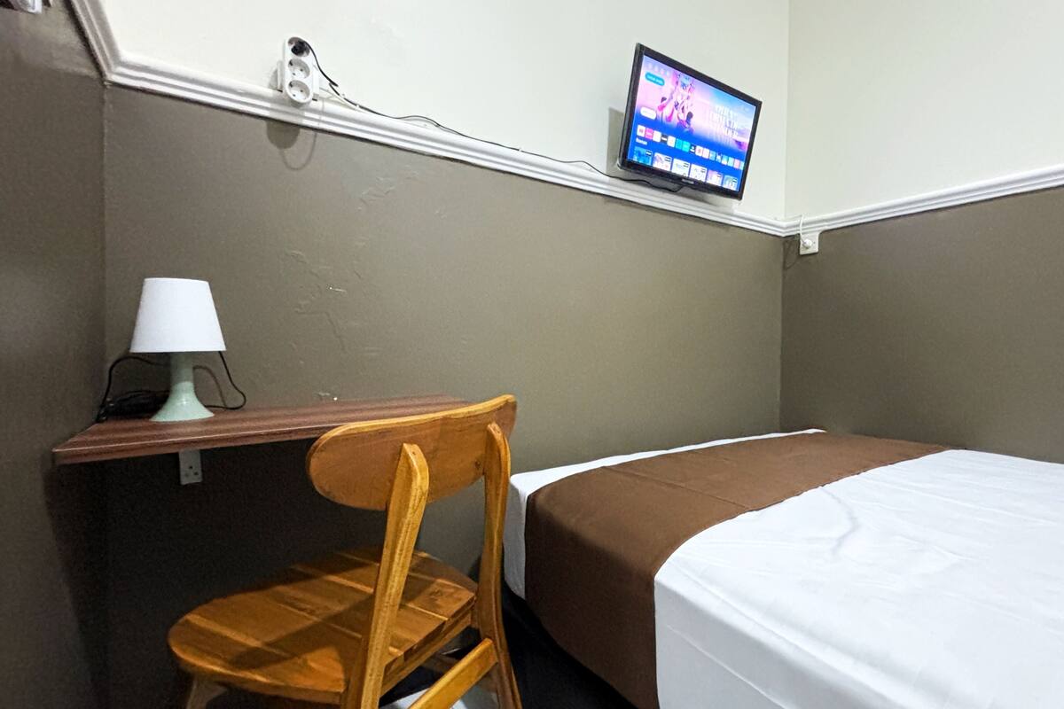 A small hotel room with a single bed, a wooden chair, a desk with a lamp, and a television mounted on the wall.