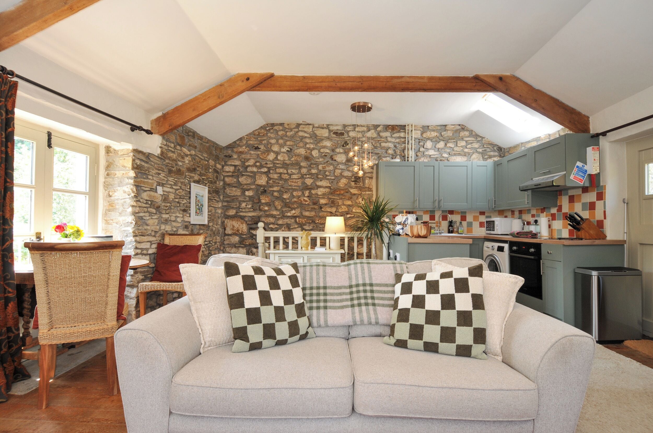 A cozy living room with a stone wall, a sofa with checkered cushions, a dining table, and a kitchen area with cabinets and appliances.