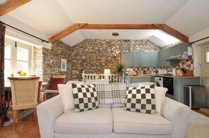 A cozy living room with a stone wall, a sofa with checkered cushions, a dining table, and a kitchen area with cabinets and appliances. - The Mill (Lostwithiel)