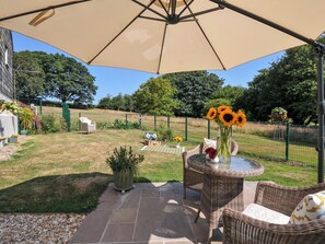 An outdoor patio area with a covered seating area, a table with a vase of sunflowers, and a view of a fenced garden. - The Mill (Lostwithiel)