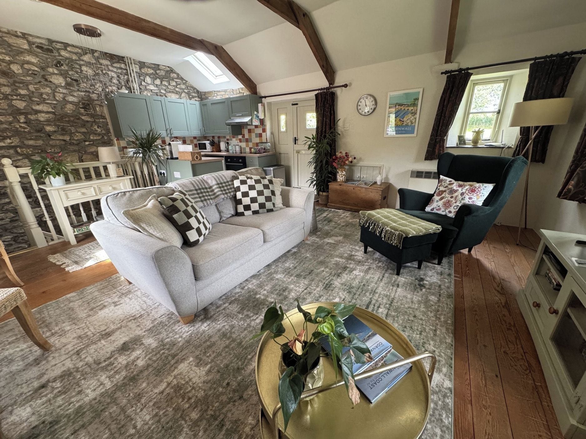 A cozy living room with a sofa, armchairs, and a coffee table. There is a kitchen in the background with a stone wall and wooden beams.