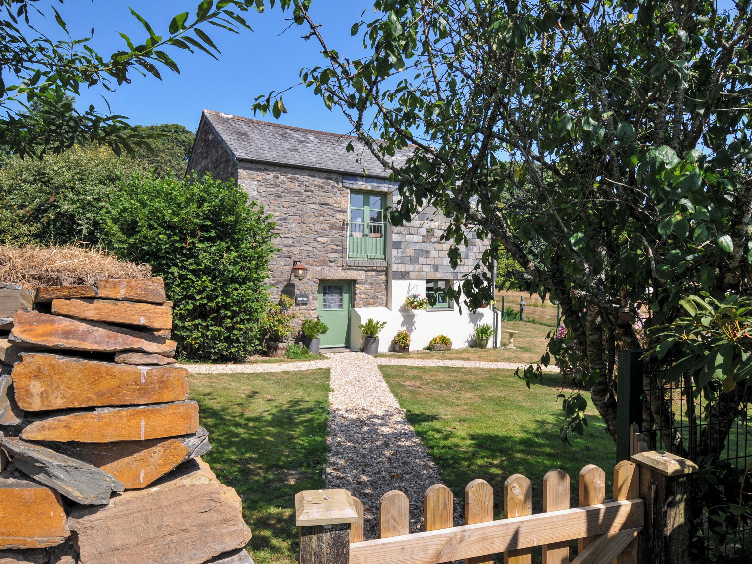 Ein Steinhaus mit grüner Tür, ein Kiesweg, der zum Eingang führt, und ein Holzzaun im Vordergrund.
