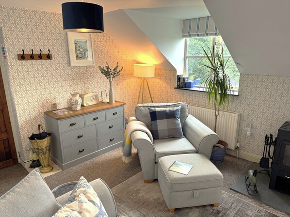 A cozy living room with a sofa, a side table, a lamp, a radiator, and a window with a view of greenery.