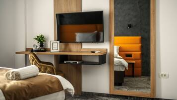 Quarto de hotel moderno com cama, escrivaninha com televisão de tela plana, uma cadeira, uma pequena planta e um espelho refletindo o ambiente.