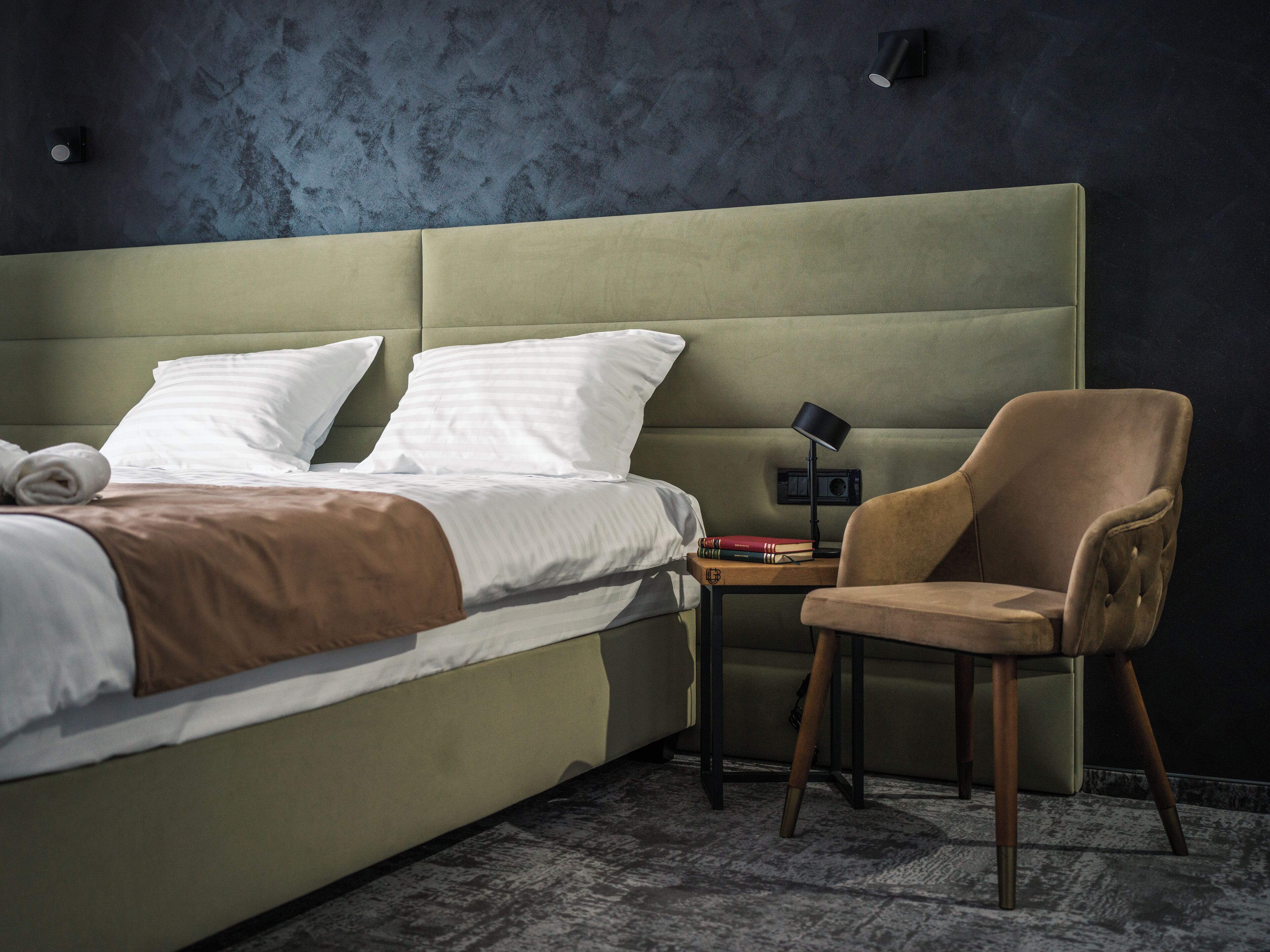 A modern hotel room with a large bed, a wooden chair, and a small table with a lamp.
