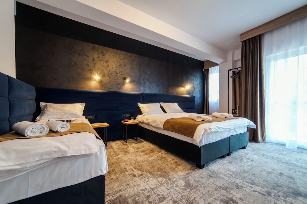 Una habitación de hotel moderna con dos camas, un cabecero grande y oscuro, y un ventanal con cortinas.