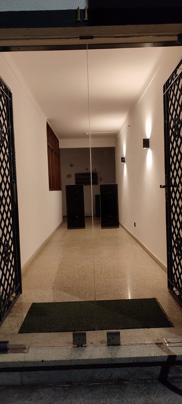 Un pasillo con alfombra que conduce a una puerta, un felpudo en la entrada y una lámpara en la pared.