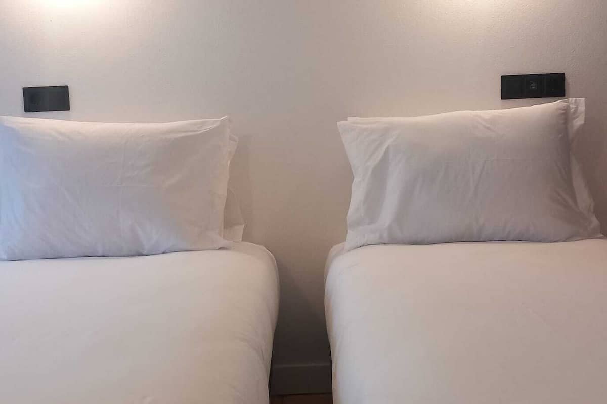 Two single beds with white linens and headboards, each with a small black control panel on the wall above.