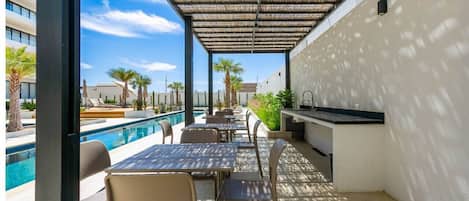 An outdoor pool area with tables and chairs, a pergola, and a kitchen area.