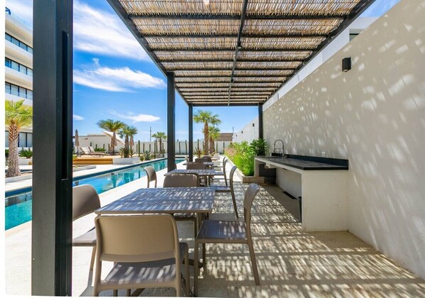 Zona de piscina exterior con mesas y sillas, una pérgola y una cocina.