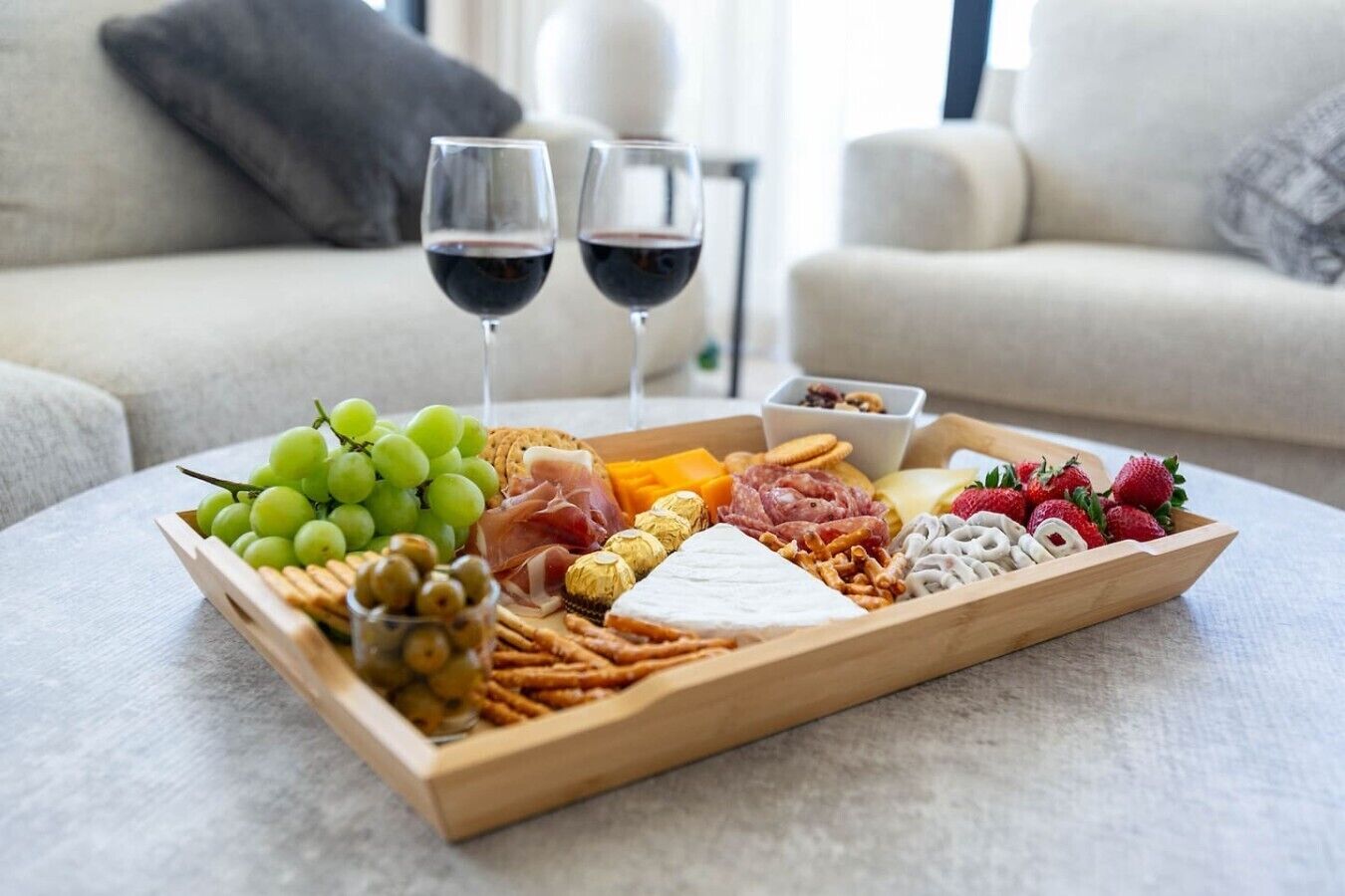 A charcuterie board with various cheeses, meats, fruits, and crackers on a tray, accompanied by two glasses of red wine.