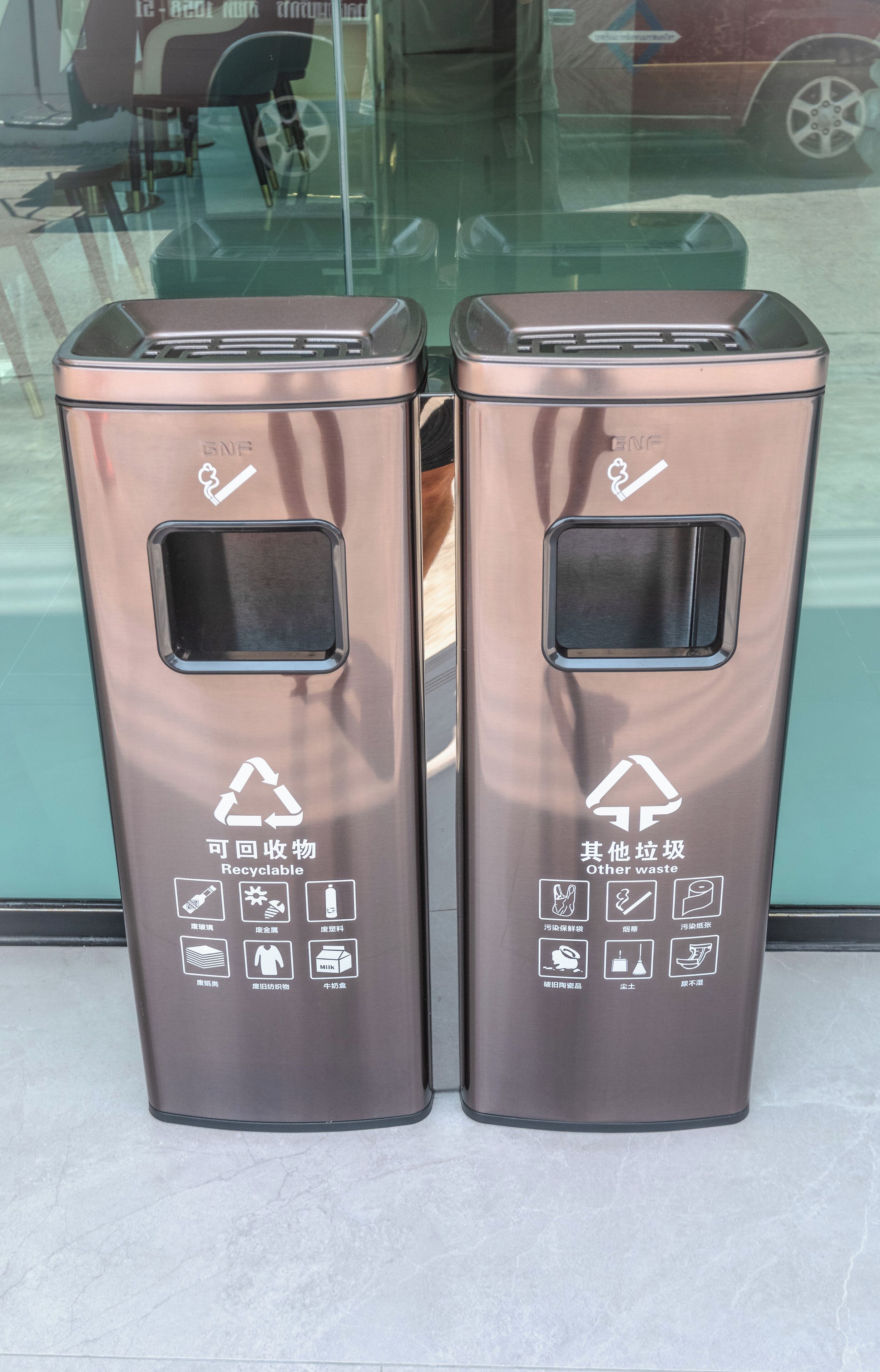 Zwei metallische Mülleimer mit Recycling- und Abfallsymbolen auf einem gefliesten Boden.