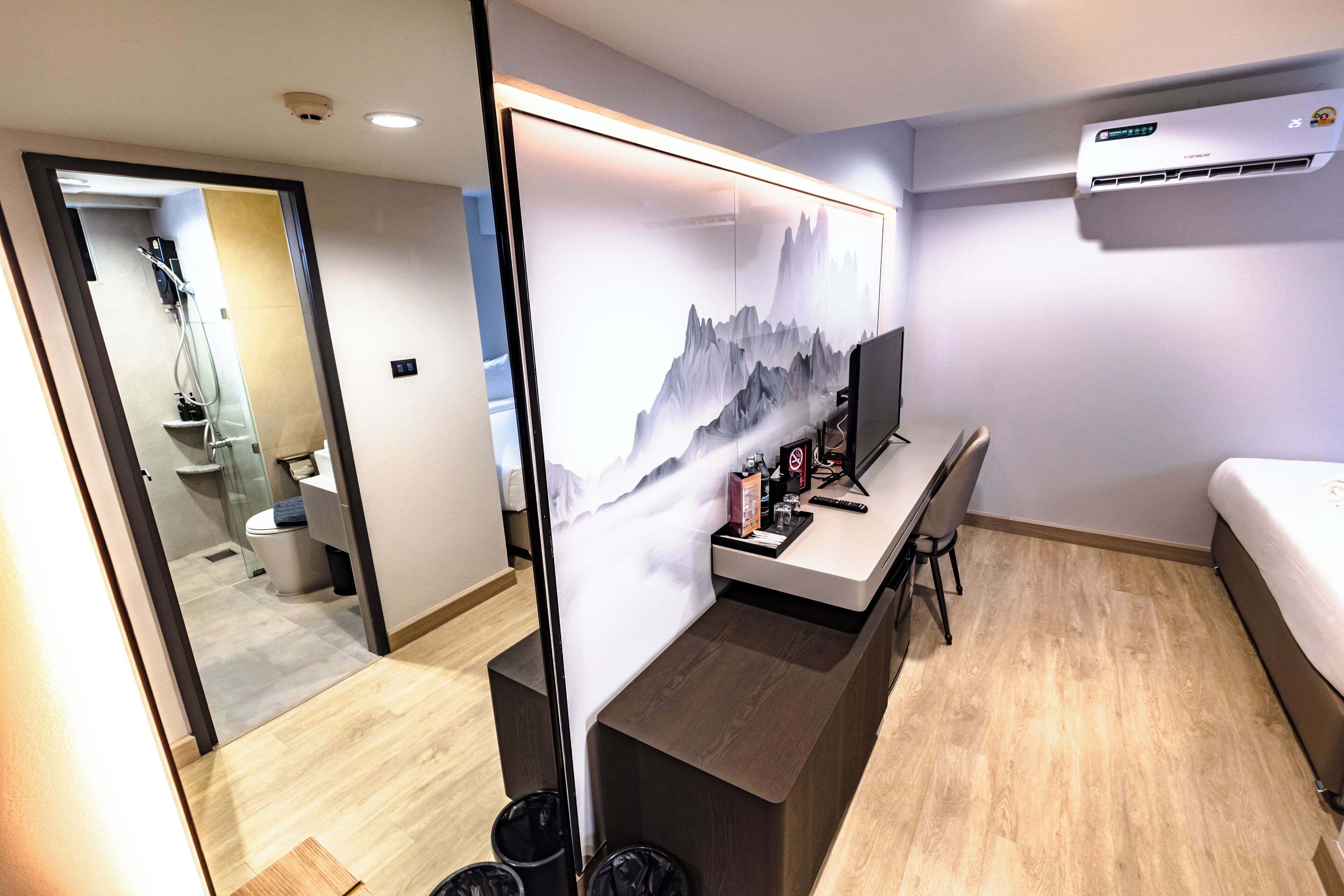 A hotel room with a wooden floor, a bed, a desk with a chair, a large mirror, and a wall-mounted air conditioner.