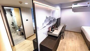 A hotel room with a wooden floor, a bed, a desk with a chair, a large mirror, and a wall-mounted air conditioner.