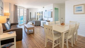 Ein modernes Wohnzimmer mit einer Couch, einem Esstisch mit Stühlen, einem Fernseher und einem großen Fenster mit Vorhängen.
