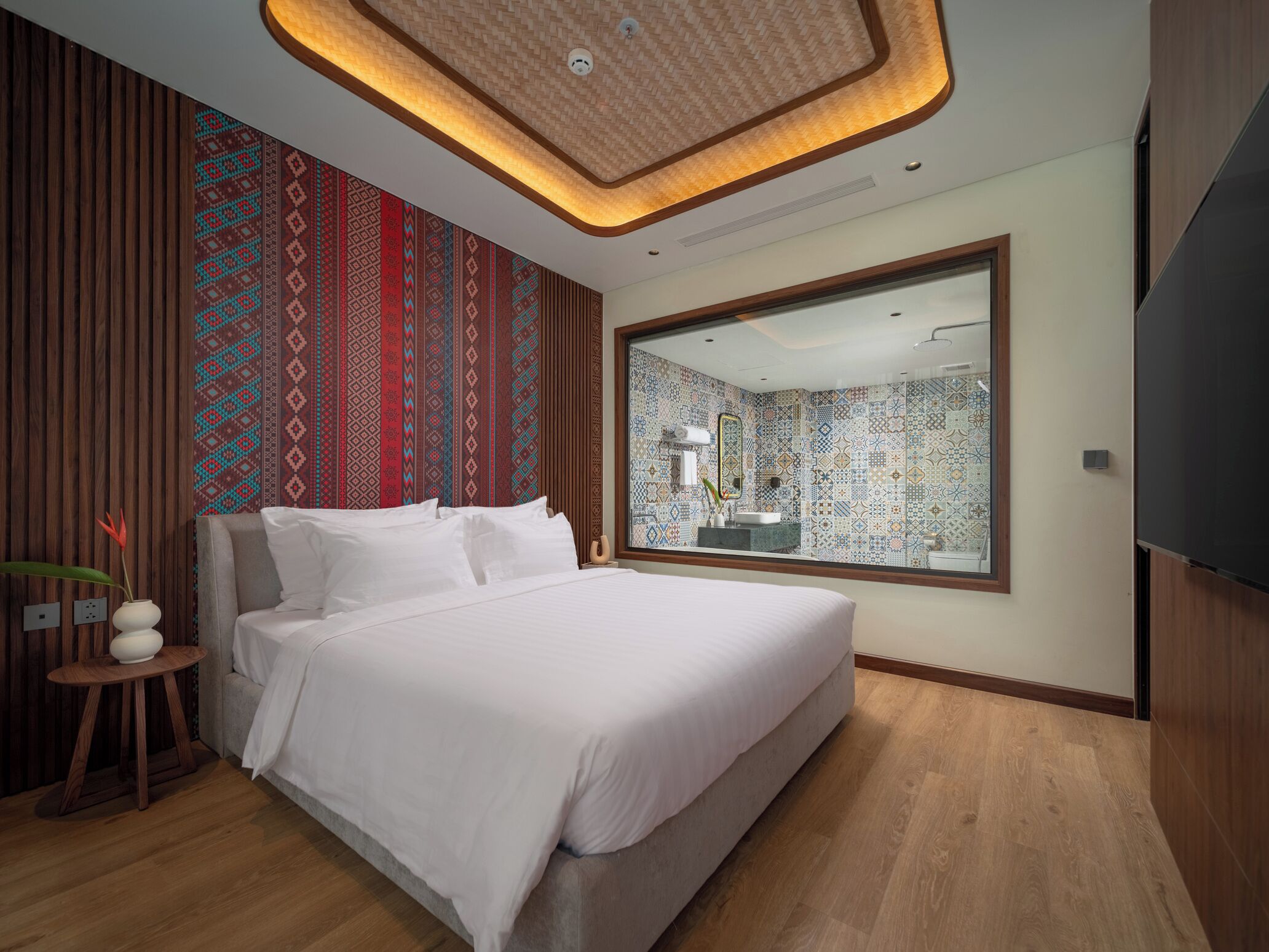 Een moderne slaapkamer met een groot bed, een houten vloer, een behang met patroon en een grote spiegel die de badkamer weerspiegelt.
