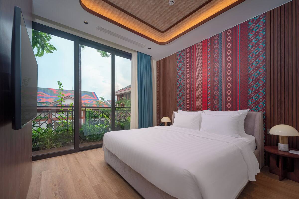 Habitación de hotel moderna con una cama grande, un ventilador de techo y vistas a un edificio colorido a través de la ventana.