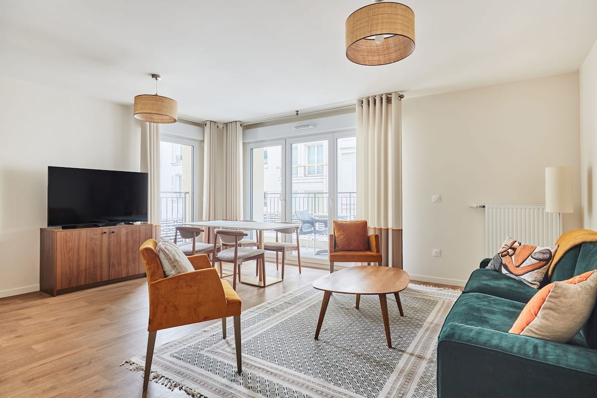 A modern living room with a flat-screen TV, wooden furniture, a green sofa, and large windows.