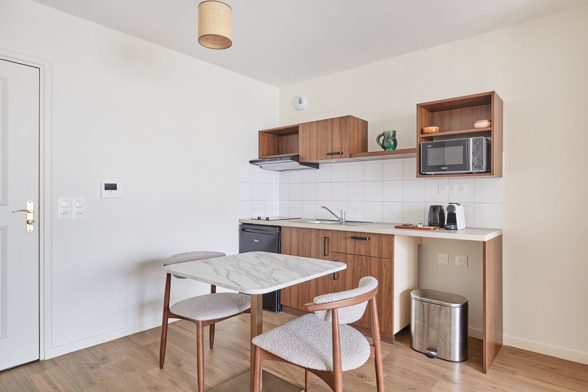 A compact kitchen with a white marble table, two wooden chairs, a microwave, a coffee maker, and a trash bin.