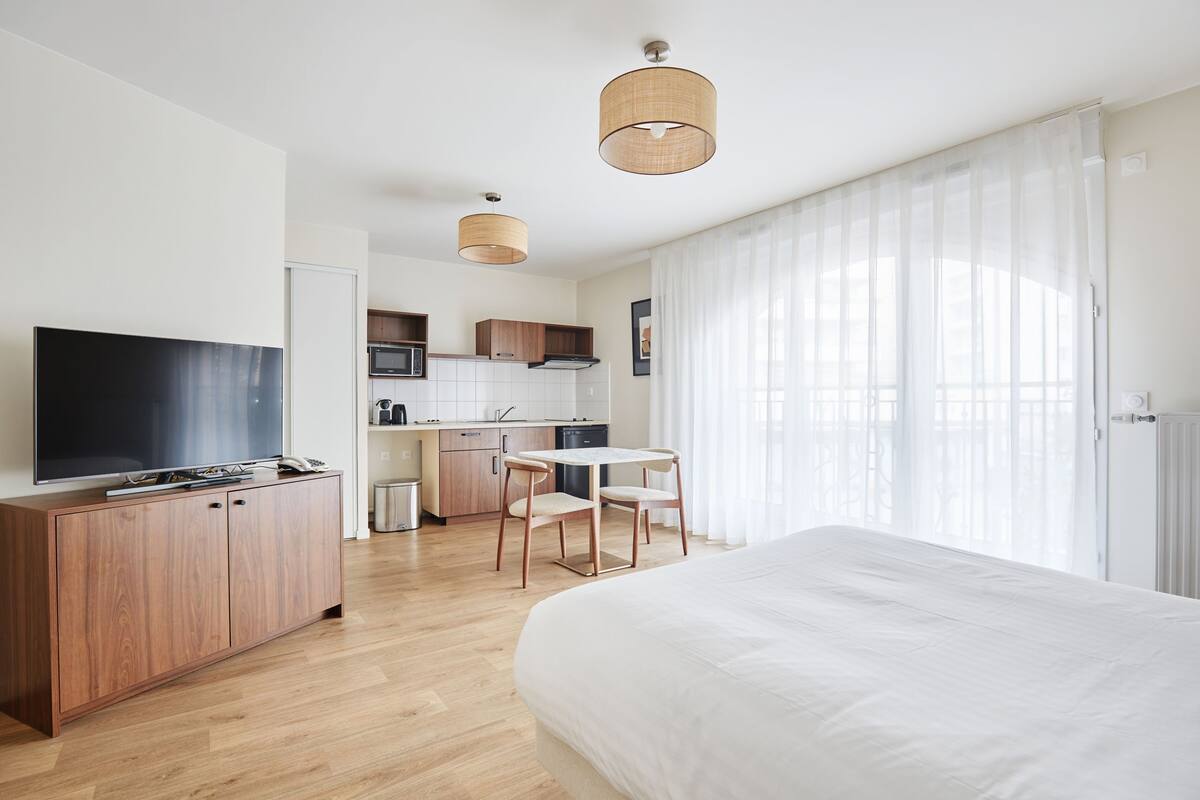 A modern hotel room with a large bed, a TV, a wooden cabinet, a kitchen area, and a dining table.