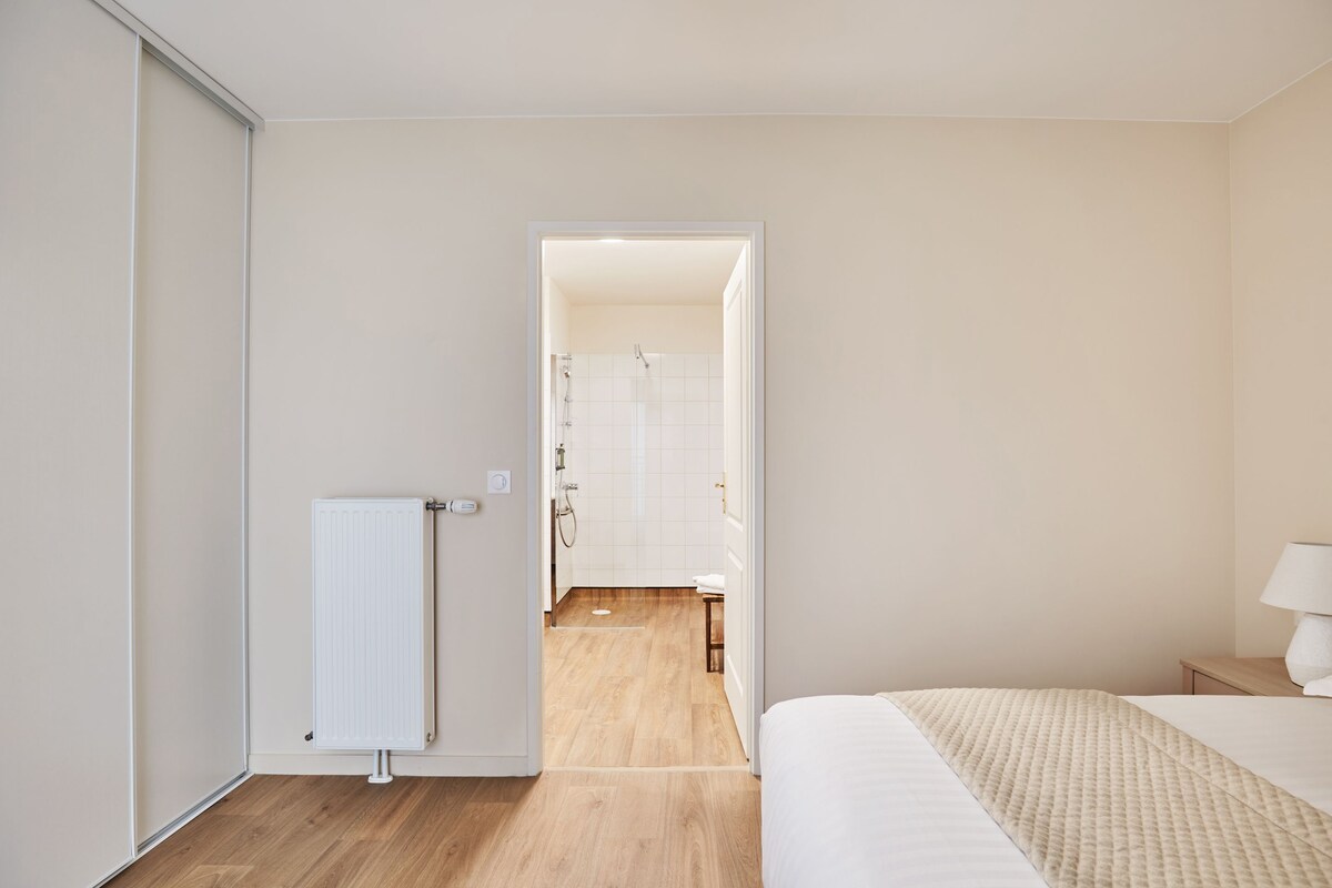 Un dormitorio con cama, mesita de noche, radiador y vista a un baño contiguo.