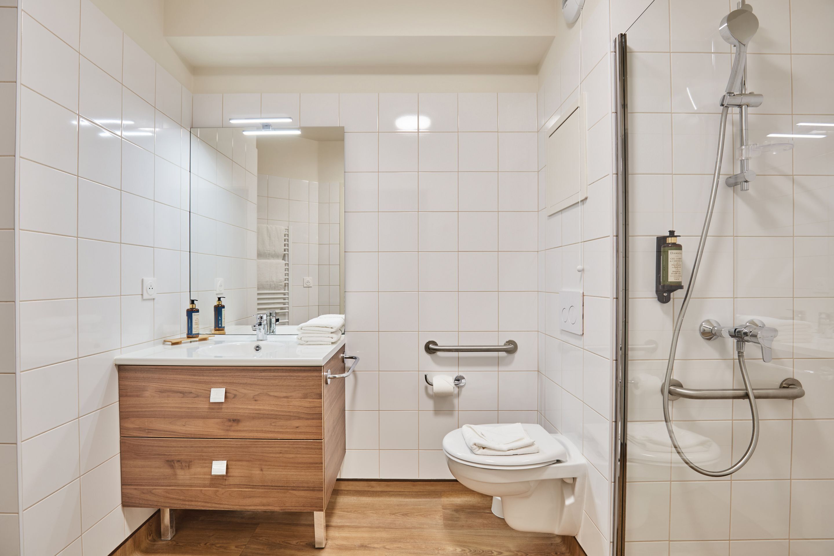 Moderni kylpyhuone, jossa on valkoiset laatat, puinen allaskaappi, wc ja suihkukaappi.